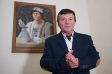 Muž v modrém obleku, za ním je obraz se stejným mužem, ale mladším, v pohádkovém převleku prince