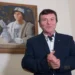 Muž v modrém obleku, za ním je obraz se stejným mužem, ale mladším, v pohádkovém převleku prince