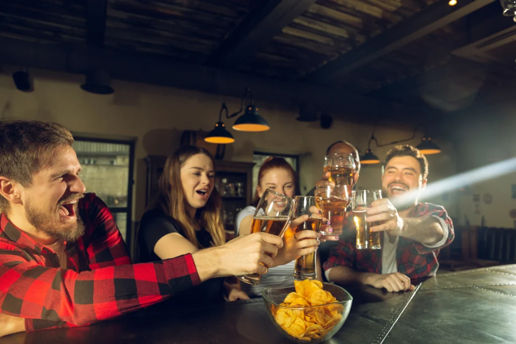 shutterstock_1670720692 - VIPshow Skupina mladých lidí v hodspodě si připíjí pivem, muž v popředí má červenou flanelovou košili, žena má dlouhé vlasy a otevřená ústa
