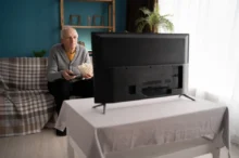 Starý muž sedí na gauči pokrytém károvanou dekou a dívá se na televizi. Jí popcorn