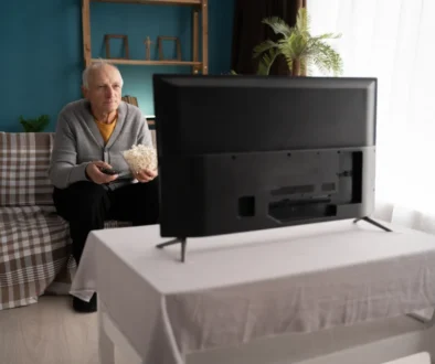 Starý muž sedí na gauči pokrytém károvanou dekou a dívá se na televizi. Jí popcorn