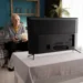 Starý muž sedí na gauči pokrytém károvanou dekou a dívá se na televizi. Jí popcorn