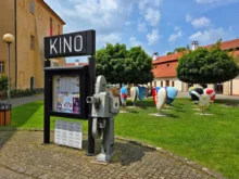 Louka před kinem, na ní je cedule s nápisem Kino a několik plastik ve tvaru srdce s různými motivy
