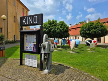 Louka před kinem, na ní je cedule s nápisem Kino a několik plastik ve tvaru srdce s různými motivy
