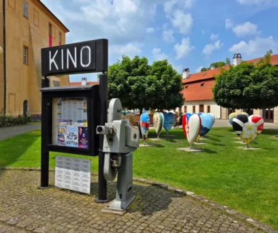 Louka před kinem, na ní je cedule s nápisem Kino a několik plastik ve tvaru srdce s různými motivy