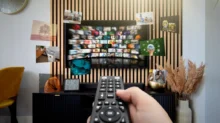 Ruka drží ovladač namířený na televizní obrazovku, na ní jsou desítky kostiček s různými televizními stanicemi