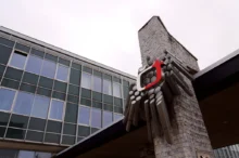 Low-angle view of a modern glass-fronted building with a stone tower and a red sculptural accent