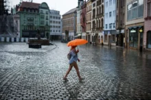 Person walking across a rainy cobblestone square with an orange umbrella, colorful buildings in the background.