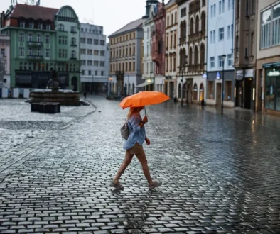 Person walking across a rainy cobblestone square with an orange umbrella, colorful buildings in the background.