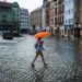 Person walking across a rainy cobblestone square with an orange umbrella, colorful buildings in the background.