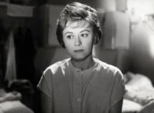 Black-and-white photo of a woman with short hair and a buttoned blouse, looking off to the side in a dim indoor setting.