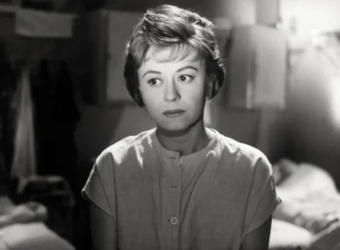 Black-and-white photo of a woman with short hair and a buttoned blouse, looking off to the side in a dim indoor setting.