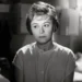 Black-and-white photo of a woman with short hair and a buttoned blouse, looking off to the side in a dim indoor setting.