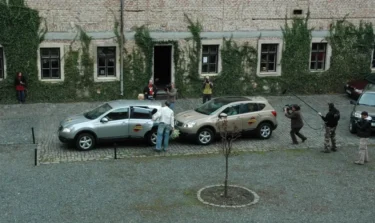 Film crew photographing a scene with two SUVs parked on a cobblestone street in front of a ivy-covered brick building, a man hands something to a passenger in the silver car.