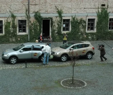 Film crew photographing a scene with two SUVs parked on a cobblestone street in front of a ivy-covered brick building, a man hands something to a passenger in the silver car.