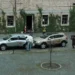 Film crew photographing a scene with two SUVs parked on a cobblestone street in front of a ivy-covered brick building, a man hands something to a passenger in the silver car.