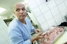 Muž bez vlasů stojí v kuchyni a sklání se nad tác s masem