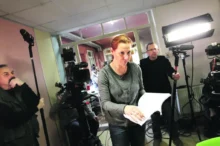 Žena v tmavém tričku a se zrzavými vlasy drží v ruce svázané papíry a listuje v nich