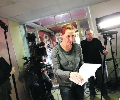 Žena v tmavém tričku a se zrzavými vlasy drží v ruce svázané papíry a listuje v nich