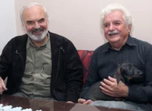 Dva šedovlasí muži sedí za stolem, jeden drží psa, oba se usmívají