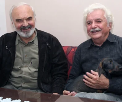 Dva šedovlasí muži sedí za stolem, jeden drží psa, oba se usmívají