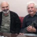Dva šedovlasí muži sedí za stolem, jeden drží psa, oba se usmívají