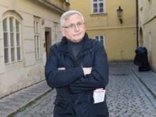 Starší muž s šedivými vlasy stojí v tmavém kabátě na ulici a ruce má zkřížené na prsou