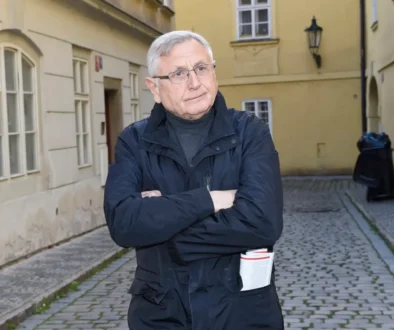 Starší muž s šedivými vlasy stojí v tmavém kabátě na ulici a ruce má zkřížené na prsou
