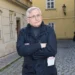 Starší muž s šedivými vlasy stojí v tmavém kabátě na ulici a ruce má zkřížené na prsou