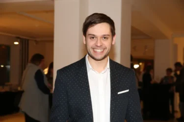 Mladý muž s tmavými vlasy, v tmavém saku a bílé košili se usmívá do kamery
