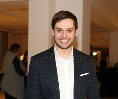 Mladý muž s tmavými vlasy, v tmavém saku a bílé košili se usmívá do kamery