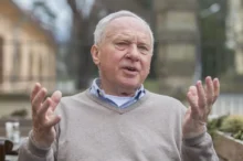 Starší muž s šedivými vlasy sedí venku a gestikuluje