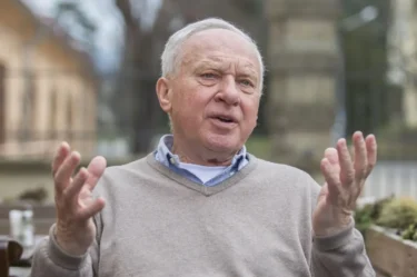 Starší muž s šedivými vlasy sedí venku a gestikuluje