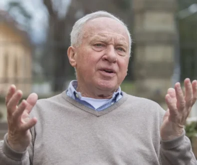 Starší muž s šedivými vlasy sedí venku a gestikuluje