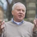 Starší muž s šedivými vlasy sedí venku a gestikuluje