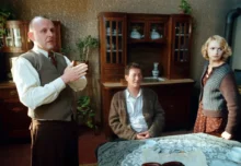 Dva muži a jedna žena jsou u jednoho stolu v dobových kostýmech a za nimi je stará komoda