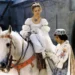 Princess in a silver puff-sleeve gown sits sidesaddle on a white horse, wearing a tiara and blue pendant, as a costumed attendant helps her.