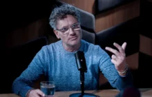 Man in a blue knit sweater speaks into a microphone during a podcast, gesturing with his right hand while seated at a wooden table with a glass of water nearby