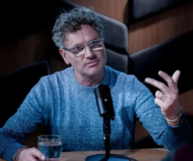 Man in a blue knit sweater speaks into a microphone during a podcast, gesturing with his right hand while seated at a wooden table with a glass of water nearby