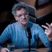 Man in a blue knit sweater speaks into a microphone during a podcast, gesturing with his right hand while seated at a wooden table with a glass of water nearby