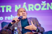 Starší muž bez vlasů sedí v křesle a mluví do mikrofonu