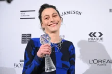 Žena ve večerních šatech stojí před zdí s logy se skleněnou cenou v ruce