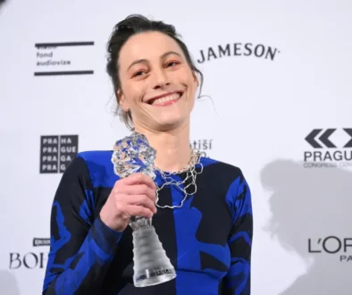 Žena ve večerních šatech stojí před zdí s logy se skleněnou cenou v ruce