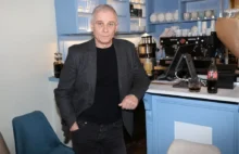 Muž v tmavém triku, tmavém saku a kalhotách stojí opřený o bledě modrý pult