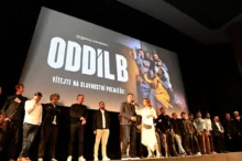 Lidé stojí na podiu před velkým plátnem, na kterém se zobrazuje upoutávka na nový seriál