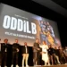 Lidé stojí na podiu před velkým plátnem, na kterém se zobrazuje upoutávka na nový seriál