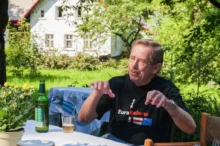Starší muž v černém tričku sedí na židli na zahradě a gestikuluje