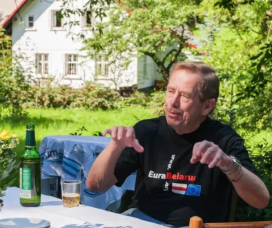 Starší muž v černém tričku sedí na židli na zahradě a gestikuluje