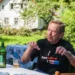 Starší muž v černém tričku sedí na židli na zahradě a gestikuluje