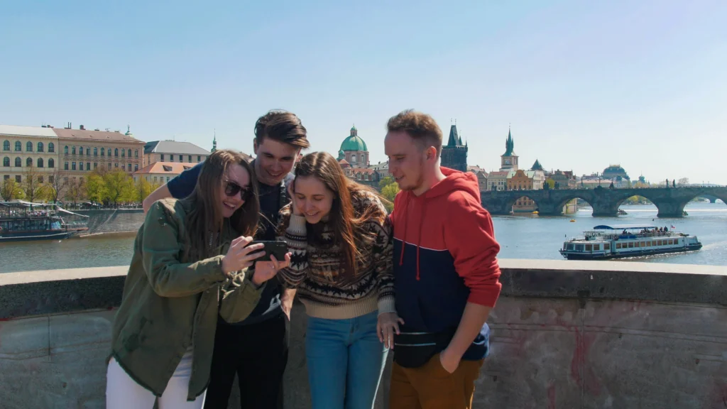 Skupina lidí v podzimním oblečení na Karlově mostě v Praze si prohlíží v mobilu fotky